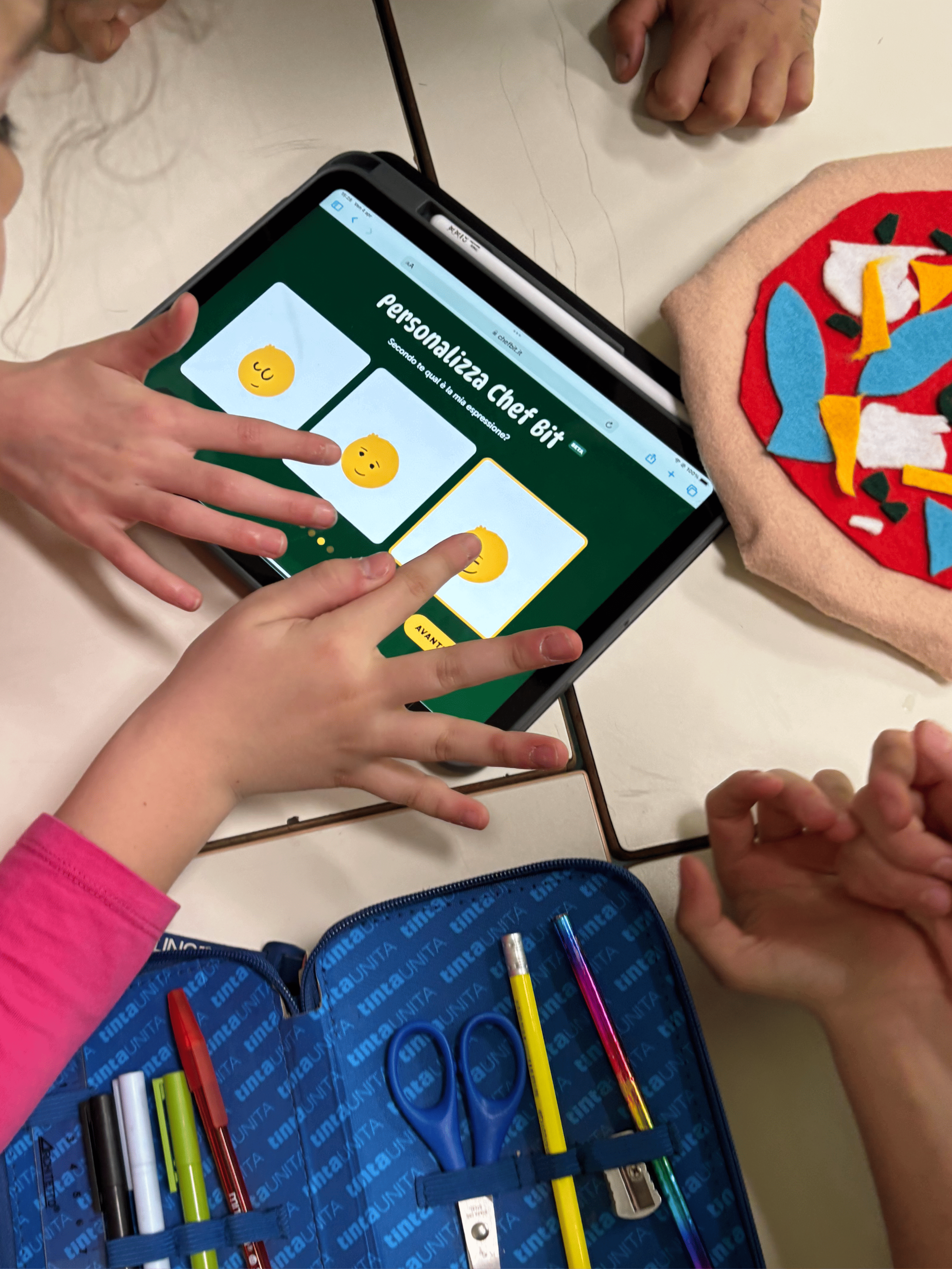 Bambini interagiscono con un tablet scegliendo un avatar su “Personalizza Chef Bit”, accanto a una pizza di feltro e un astuccio con cancelleria.