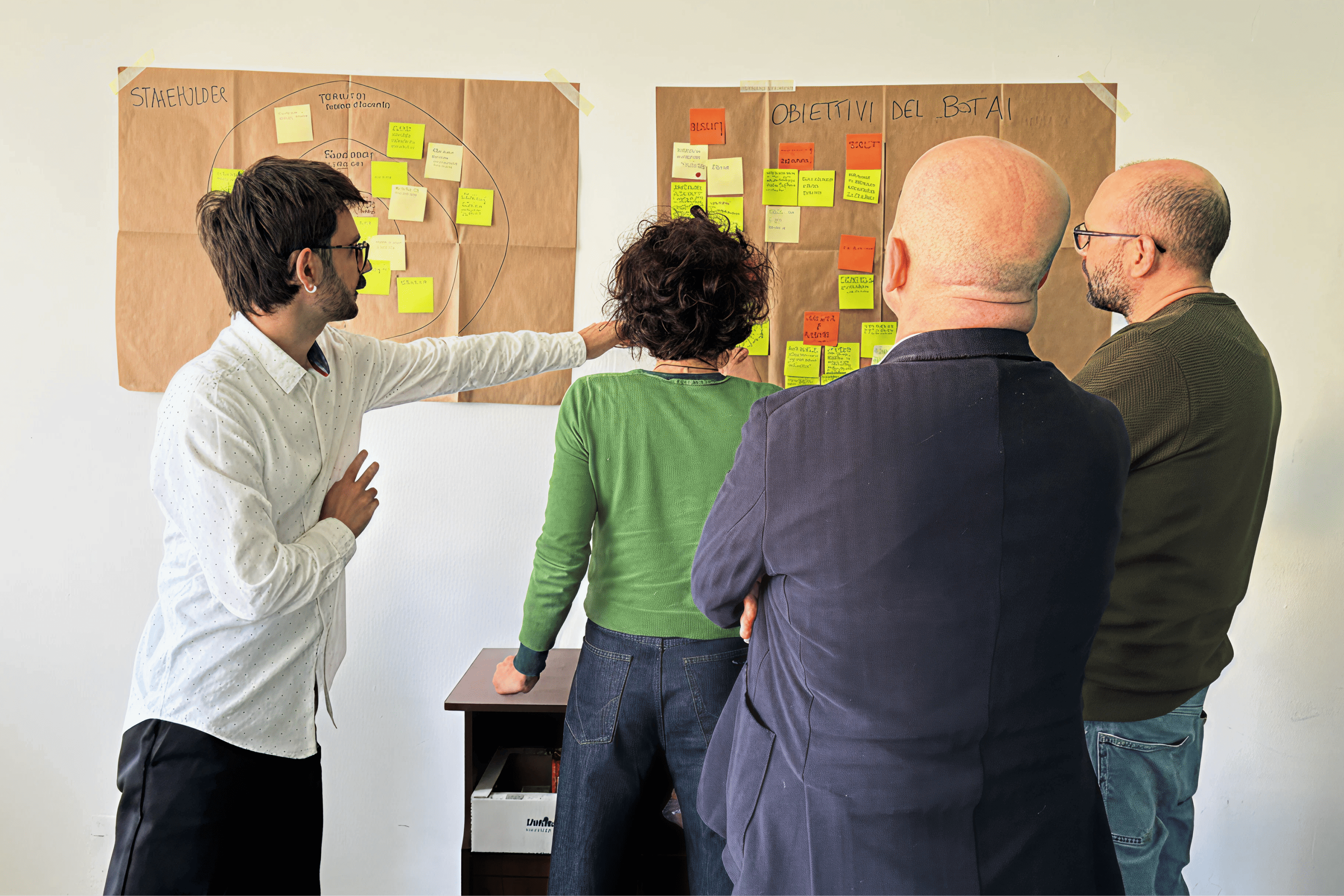 Quattro persone osservano e discutono due pannelli a muro pieni di post-it colorati, lavorando a obiettivi e stakeholder in una sessione collaborativa.
