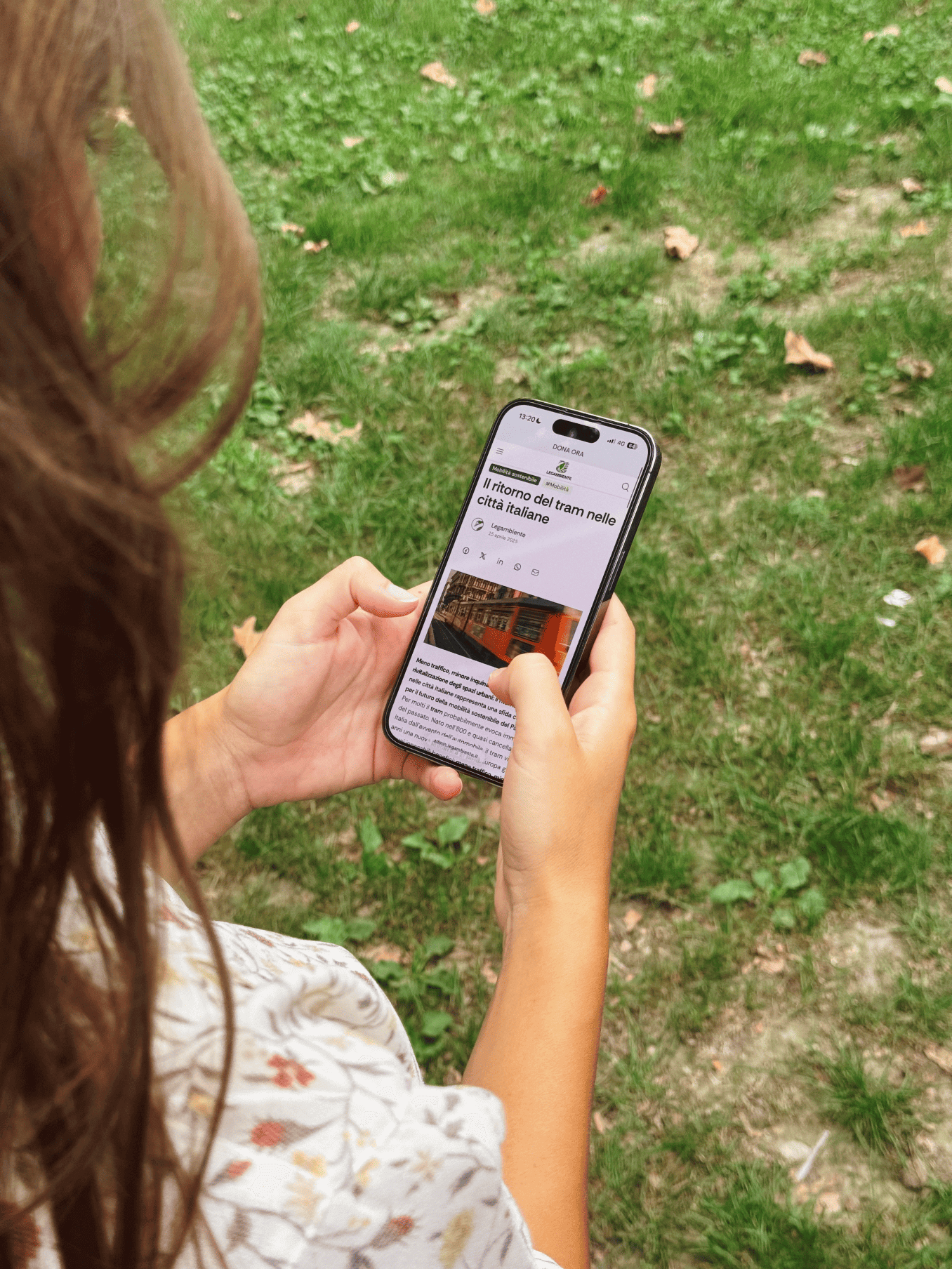 Donna all’aperto tiene in mano uno smartphone che mostra un articolo intitolato ‘Il ritorno del tram nelle città italiane’, con prato verde sullo sfondo.