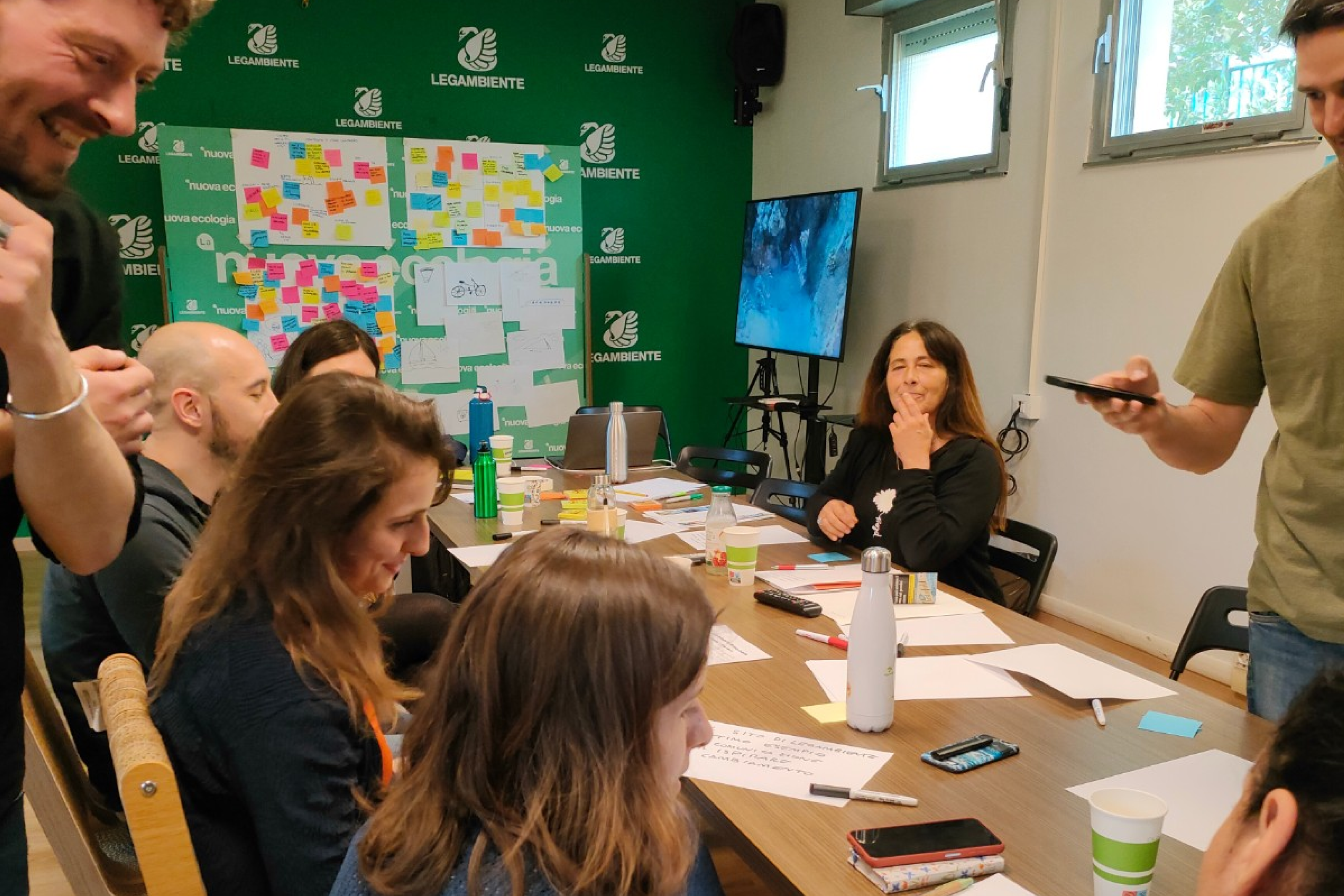 Gruppo di persone sedute a un tavolo durante un workshop con fogli, penne e post-it colorati; sullo sfondo pannelli con note e logo Legambiente.