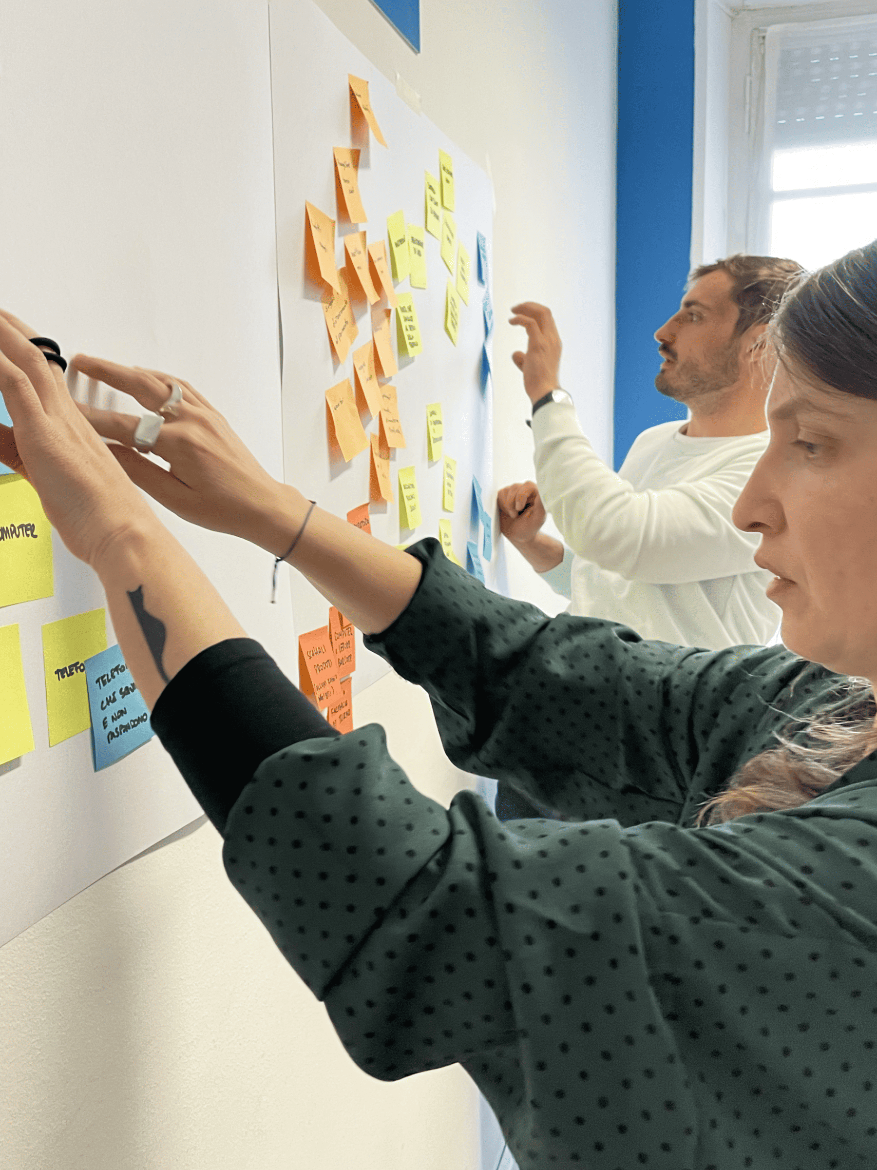 Due persone applicano e organizzano post-it colorati su una parete durante una sessione di lavoro collaborativo in ufficio, concentrati sulle idee emerse.