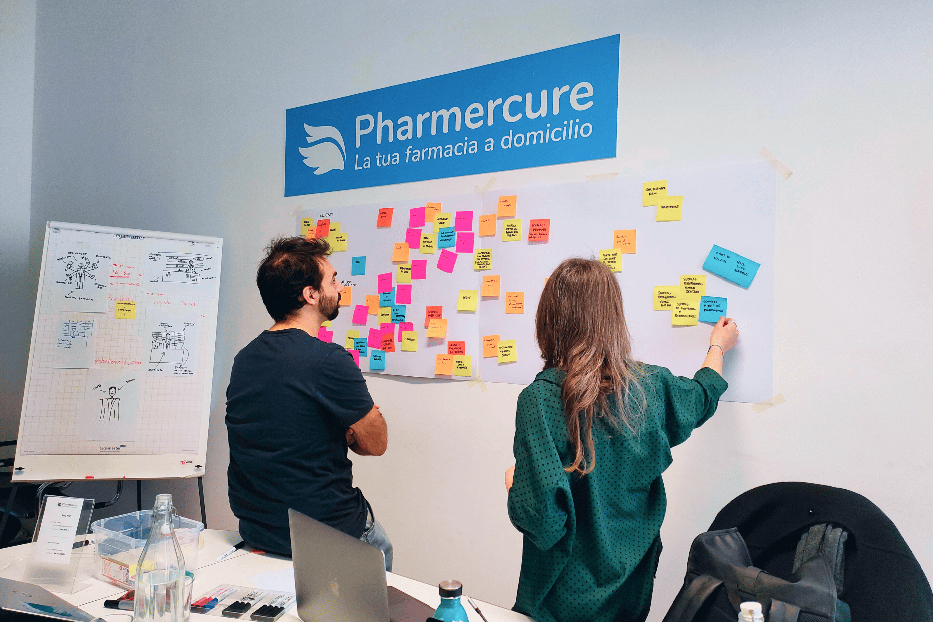 Due persone lavorano a un brainstorming davanti a un muro pieno di post-it colorati nell’ufficio Pharmercure, con lavagna a fogli mobili e materiali sul tavolo.